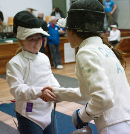Marina Carrier shakes the hand of the young fencer she has just bouted with. Both have their masks pushed back on the tops of their heads, it IS a thing! The young fencer smiles and, although we can't see Marina's face because the shot is taken from behind her and to the side, I reckon she is smiling too. She's actually such a beacon of positivity that she always seems to be smiling! Yeah, we're fans.