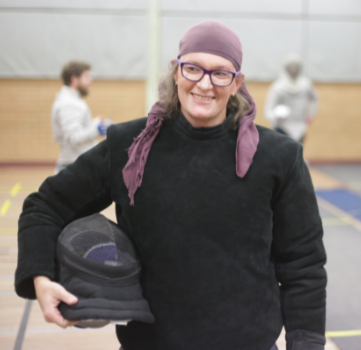 Coach Imogen Waugh, well coach Doctor Imogen Waugh actually (not that kind of doctor, but she's useful all the same), looking very cool in blacks! She's standing in the fencing hall holding her coaching mask under her right arm, wearing a purple headscarf and smiling at club founder Richard who is out of shot to the side. Behind her some sabreurs are bouting. Did you know that she can coach all three weapons both left and right handed? I know right?! What a showoff! Epee is her weapon of choice though and she's pretty damn good at it!