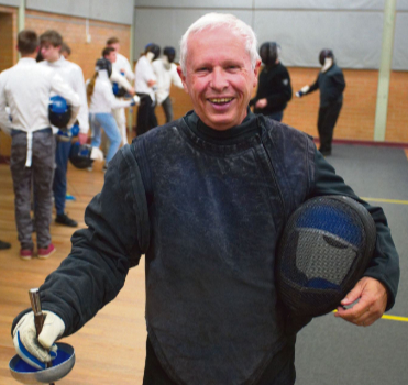Richard Emmerick resplendent in coaching blacks holding his mask under his left arm and his epee in his right hand with its point to the ground, he faces the camera smiling in the fencing hall, in the background a line of young fencers work with two of the coaches. He looks happy and why shouldn't he be? He founded the club and it's the best! 